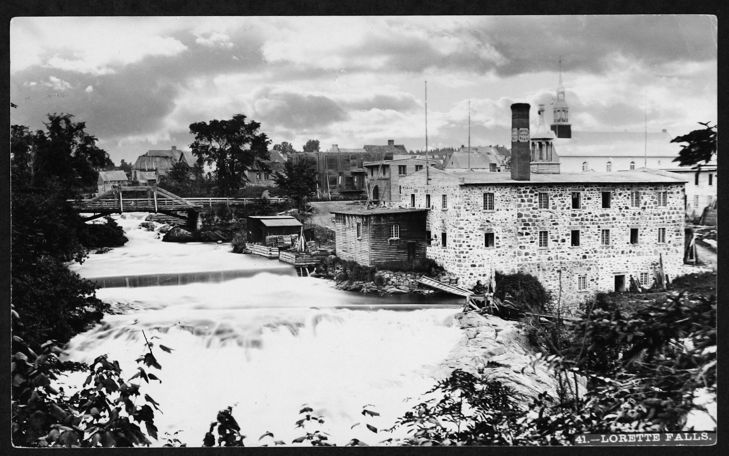Photo noir et blanc montrant un édifice en briques pâles près d'une rivière.e